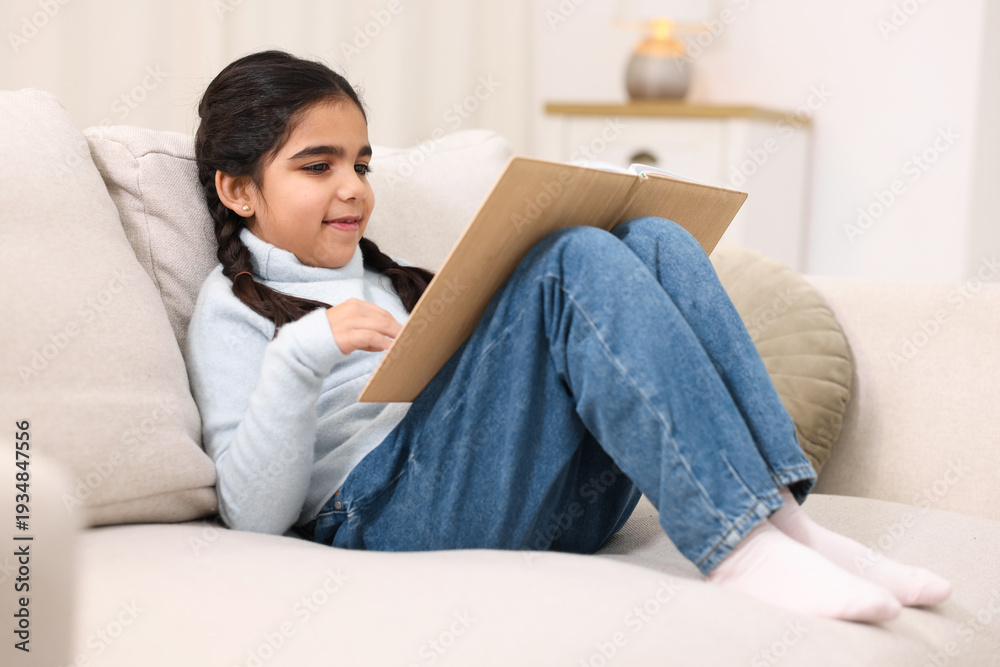 Naklejka premium Cute little girl reading book on couch at home