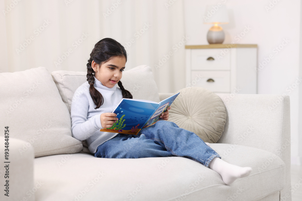 Naklejka premium Cute little girl reading book on couch at home