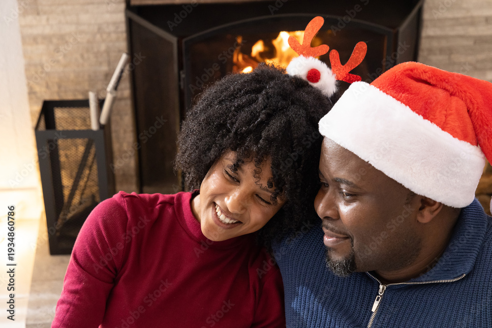 Obraz premium African American couple sitting closely at home by stone fireplace wearing Santa hat and antlers