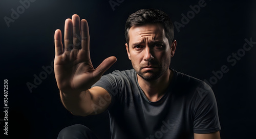 A man in a dark gray t-shirt holds up his hand in a stop gesture against a black background.