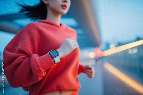East Asian Female Runner In Red Hoodie With Smartwatch, Urban Evening Jogging And Fitness Tracking Concept