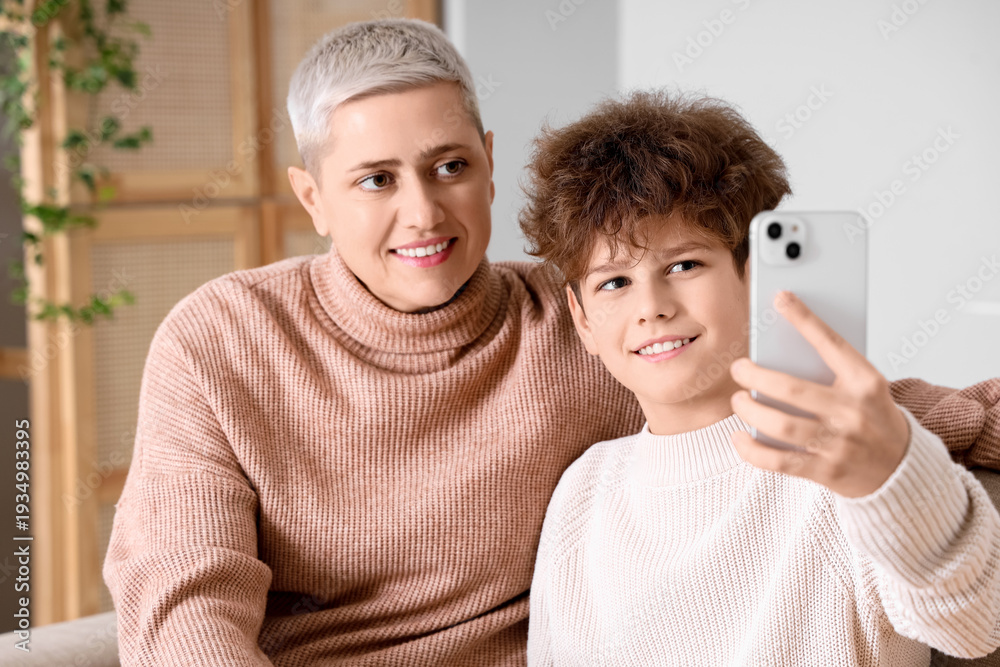 Fototapeta premium Mother and her teenage son in warm sweaters taking selfie on sofa at home