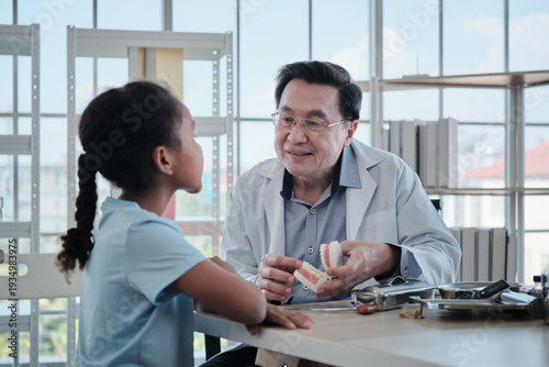 Professional senior Asian dentist explains the tooth model to Black patient girl, oral care advice, examine consultation, specialist in kids' healthcare, medical visits to pediatric clinic hospital.