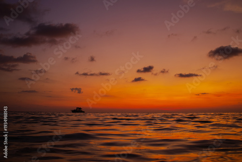 Tropical sunset seascape with dramatic orange sky and gentle waves in Thailand. Silhouette of boat on horizon, romantic travel background, calm nature scene with vibrant sunset colors.