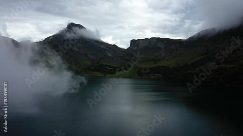 Dramatic mountain landscape with mist and clouds over a tranquil lake at Barrage de Roselend in the French Alps. Moody natural scenery under an overcast sky.