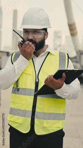 Portrait of bearded Indian contractor with clipboard talking on portable radio at construction site. Skilled manager discusses building plans against bridge columns vertical shot
