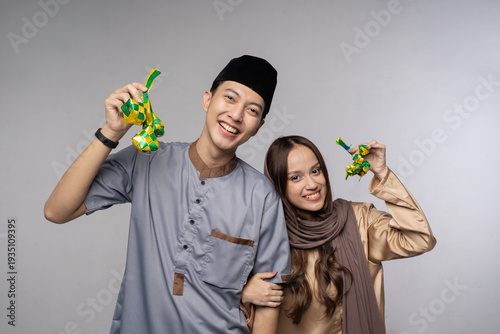 Smiling malay couple holding green envelopes in studio portrait wearing hijab and songkok, joyful giftgiving for eid celebration,