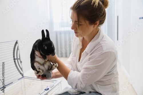 Close view of young woman holding her pet rabbit near an open cage in a bright modern apartment. Caring for a domestic animal in a calm home environment.