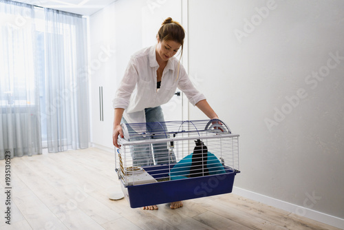 Young woman lifting and carrying a cage with her pet rabbit inside in a bright modern apartment. Daily pet care routine and domestic lifestyle.