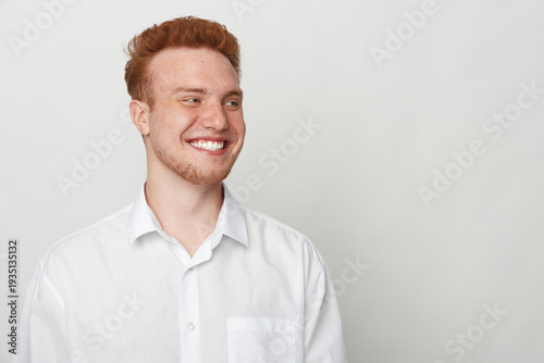 Bild auf Leinwand Smiling young man with red hair wearing white shirt looks away on light background