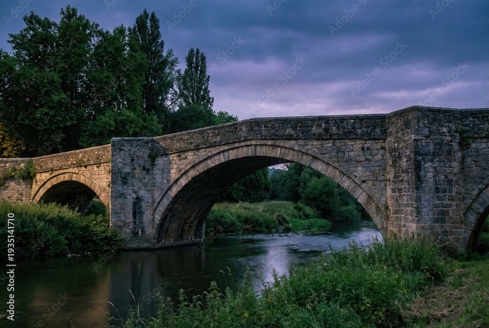 Fototapeta premium Old stone arch bridge over river at dusk with dramatic cloudy sky 
