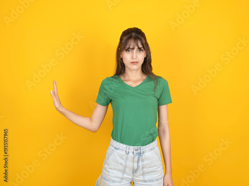 Serious young woman in green t-shirt making a stop gesture with her hand aside against a yellow background. Concept of refusal, setting boundaries, or warning.