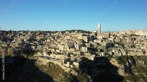 Aerial view of the historic stone buildings and dramatic cliffs of Matera, Italy, under a clear blue sky. The iconic old town and cathedral tower are visible in this panoramic landscape.