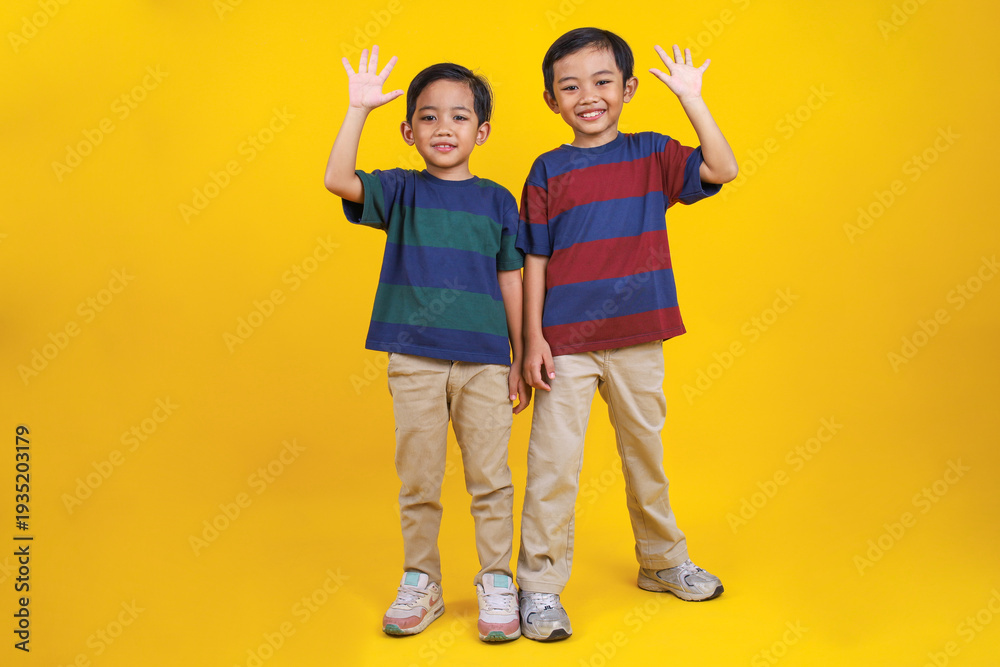 Fototapeta premium Happy and Smiling Asian Boy Stands and Waves His Hand on Yellow Background
