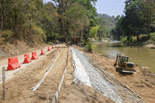 Construction site for road development and riverbank engineering. An excavator works on dirt infrastructure project next to calm river, showing progress and industry