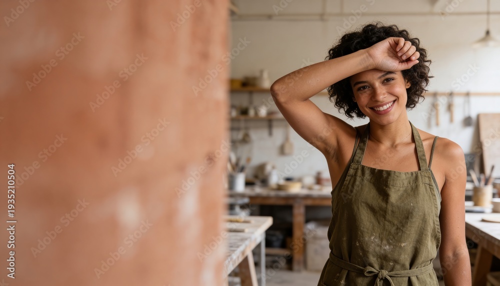 Fototapeta premium Smiling woman in linen apron relaxing in sunlit pottery studio with blurred background copy space