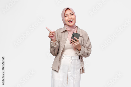Pretty young Muslim woman holding a smart phone while pointing to the copy space on her side.