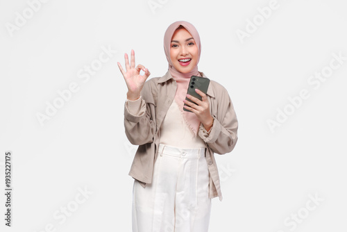 Joyful young Asian woman in hijab holding a smartphone while showing okay finger gesture.