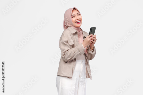 Excited young Asian Muslim woman in hijab holding smartphone with happy expression.