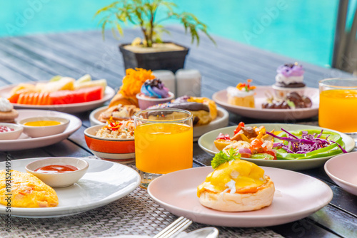Breakfast spread with fresh juice, assorted pastries, and eggs benedict, served poolside on wooden table, tropical setting.