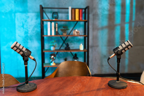Two microphones are set up on a wooden table for a podcast recording in a studio with books and decor in the background