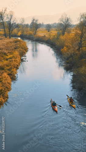 Two kayakers navigate a serene river surrounded by autumn foliage. The calm water reflects golden trees and distant hills, creating a tranquil scene.