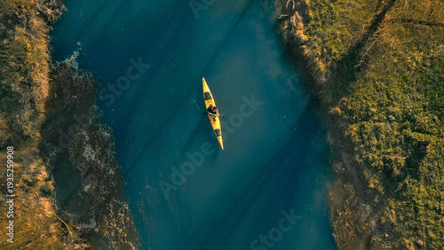 A lone kayaker navigates a serene, deep blue cove surrounded by rugged cliffs. The aerial view captures the tranquility and isolation, emphasizing the adventurous spirit of kayaking in a remote.