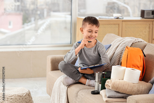 Little boy with check-list packing emergency backpack on sofa at home