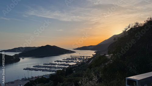 Summer evening sunset seascape over a bay near Kas, Turkiye, with yachts and ships in the harbor. The sun sets behind the mountains, and green hills surround the bay.