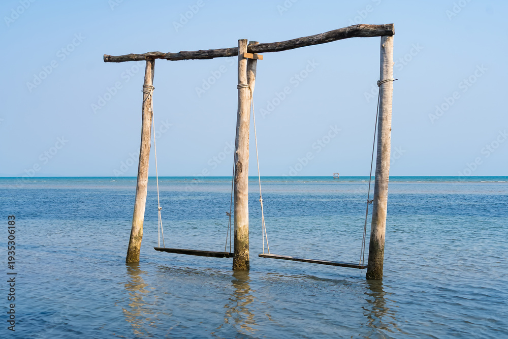 Fototapeta premium Serene Waters: A captivating beach scene features a rustic swing set emerging from the tranquil water, with the vast ocean and clear sky creating a picturesque setting.