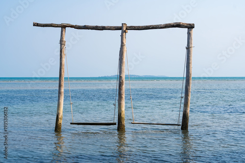 Bilde på lerret Seaside Swing Serenity: Two wooden swings stand peacefully in the tranquil shallows of the sea, inviting relaxation and offering a picturesque view of the vast ocean and clear sky