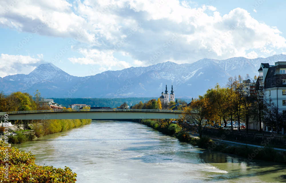 Fototapeta premium Villach, Austria - November 06, 2023: Scenic view of a river Drau with a bridge, mountains in the background, and a church tower visible in the picturesque landscape