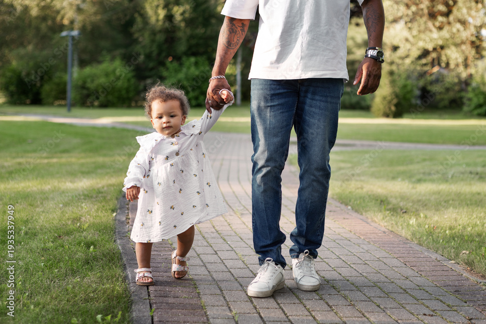 Fototapeta premium Father holding little daughter hand while child takes first steps on park path. Dad with baby outdoors