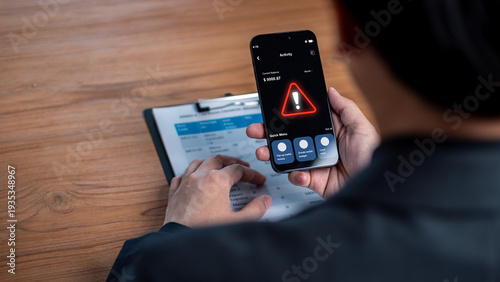 Cybersecurity risk and financial banking alert concept. Businessman holding a smartphone showing a red warning triangle icon over business documents.