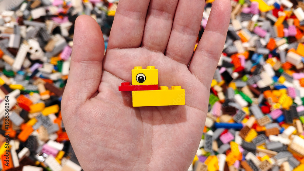 Obraz premium Small yellow and red LEGO plastic duck model held in a human hand over a background of scattered colorful building bricks on September 15, 2025 in Bangkok, Thailand.