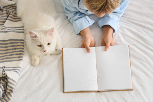 Cute little boy with cat reading book on bed, top view