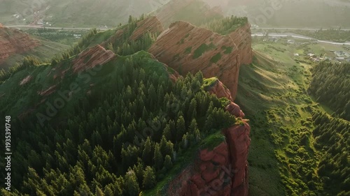 Aerial view of rugged red rock formations surrounded by lush green trees and a winding river in the valley below. Natural landscape with vibrant colors.