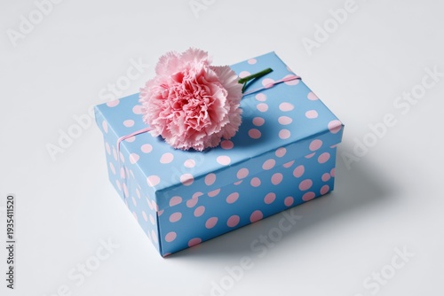 Pink carnation rests atop blue polka-dotted gift box. Soft lighting highlights delicate flower and wrapping. Minimalist background emphasizes gift and bloom