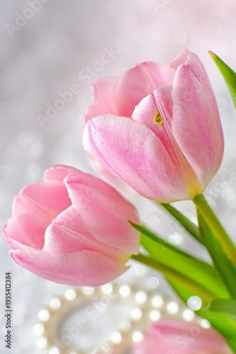 Soft pink tulips with gentle curves. Petals show subtle gradients and natural texture. Green stems add fresh, organic contrast. Pearl necklace rests softly beside blooms. Background glows with soft