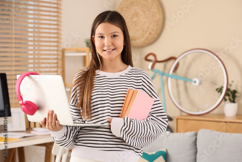 Happy teenage Asian girl with laptop, headphones and books on online educatio...