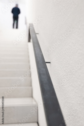 Minimalistisches weißes Treppenhaus mit Handlauf und unscharfer Person im Hintergrund.