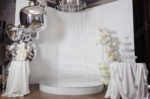 Modern luxury event backdrop featuring silver metallic balloons, a minimalist white podium, floral arrangements, and a champagne tower for gala parties