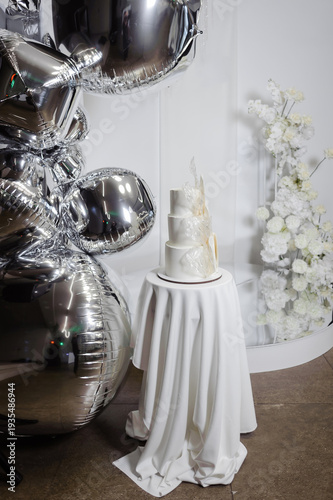 Sophisticated three-tier white wedding cake with artistic sugar decorations, displayed on a draped table with silver foil balloons and flowers.
