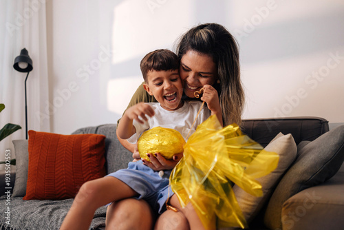 Brazilian mother surprising young son with handmade Easter egg gift on living room sofa, celebrating family affection, childhood joy and traditional holiday moments at home.