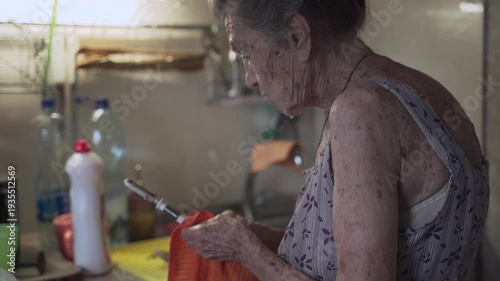 Senior elderly woman washing dishes in kitchen. Sad mature housewife cleans up the old dishes in the kitchen. Upset woman with gray hair 90 years old washing kitchen utensils in a hand wash at home.