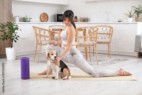 Young sporty woman with cute Beagle dog stretching on yoga mat in kitchen