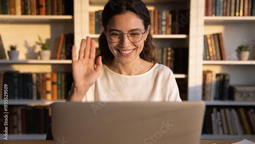 Smiling brunette woman wearing round glasses waves at her laptop screen while sitting in front of a full bookshelf illuminated by warm golden hour sunlight.