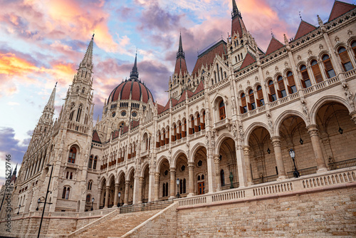The Hungarian Parliament Building stands prominently under a dramatic sunset sky with vibrant orange and purple clouds.