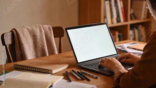 Young woman types on a slim laptop computer with a blank white screen while working at a wooden desk filled with notebooks and pens in a home office setting.