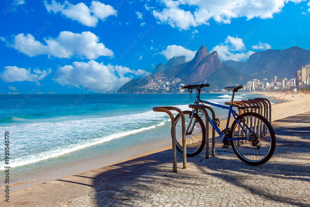 Fototapeta premium Blue mountain bike parked on the Ipanema beach boardwalk with Two Brothers Mountain (Morro Dois Irmãos) and the Atlantic Ocean in the background, Rio de Janeiro, Brazil.